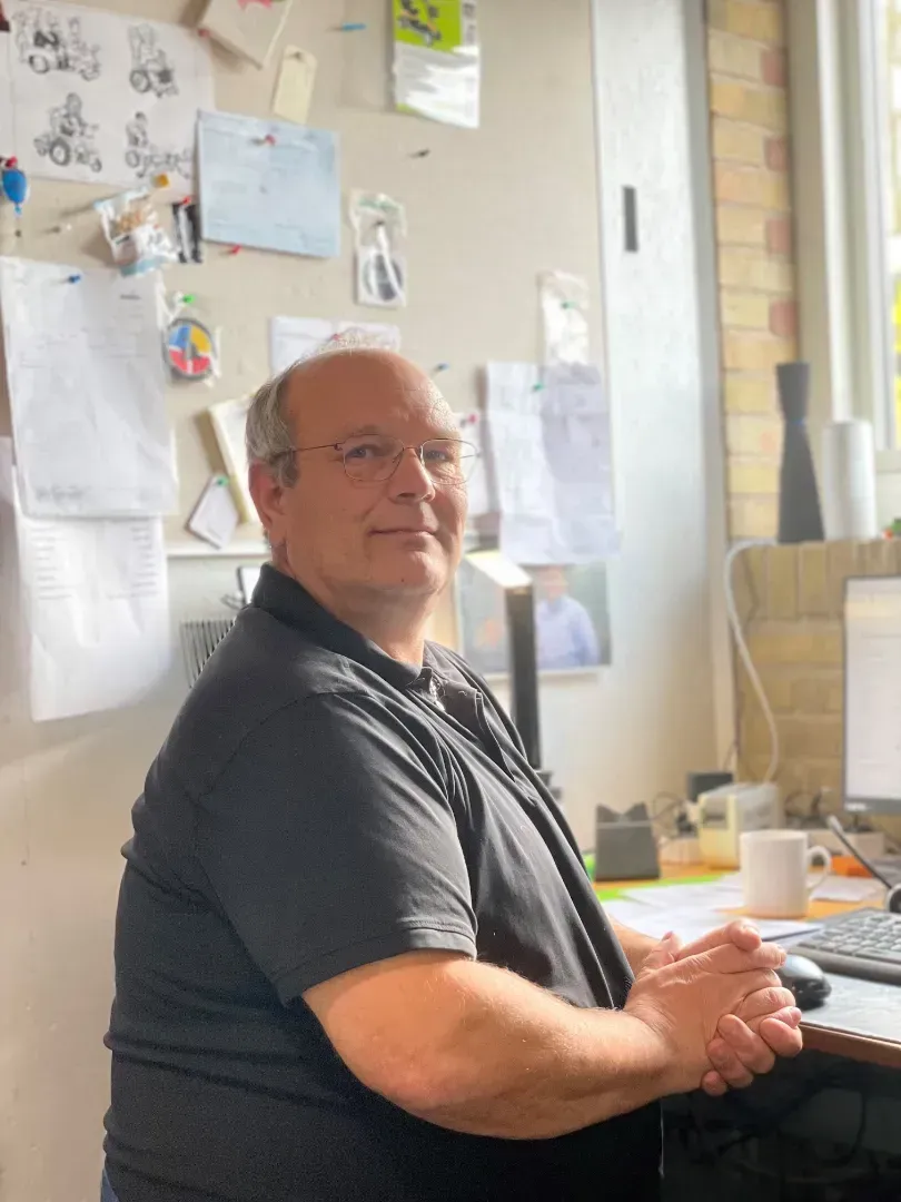 Mand med briller sidder ved et skrivebord iført en sort skjorte. Kontormiljø med papirer og en computer.