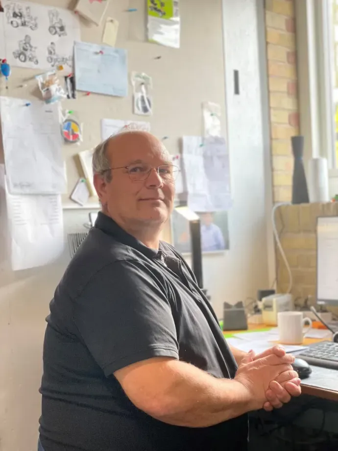 Mand med briller sidder ved et skrivebord iført en sort skjorte. Kontormiljø med papirer og en computer.