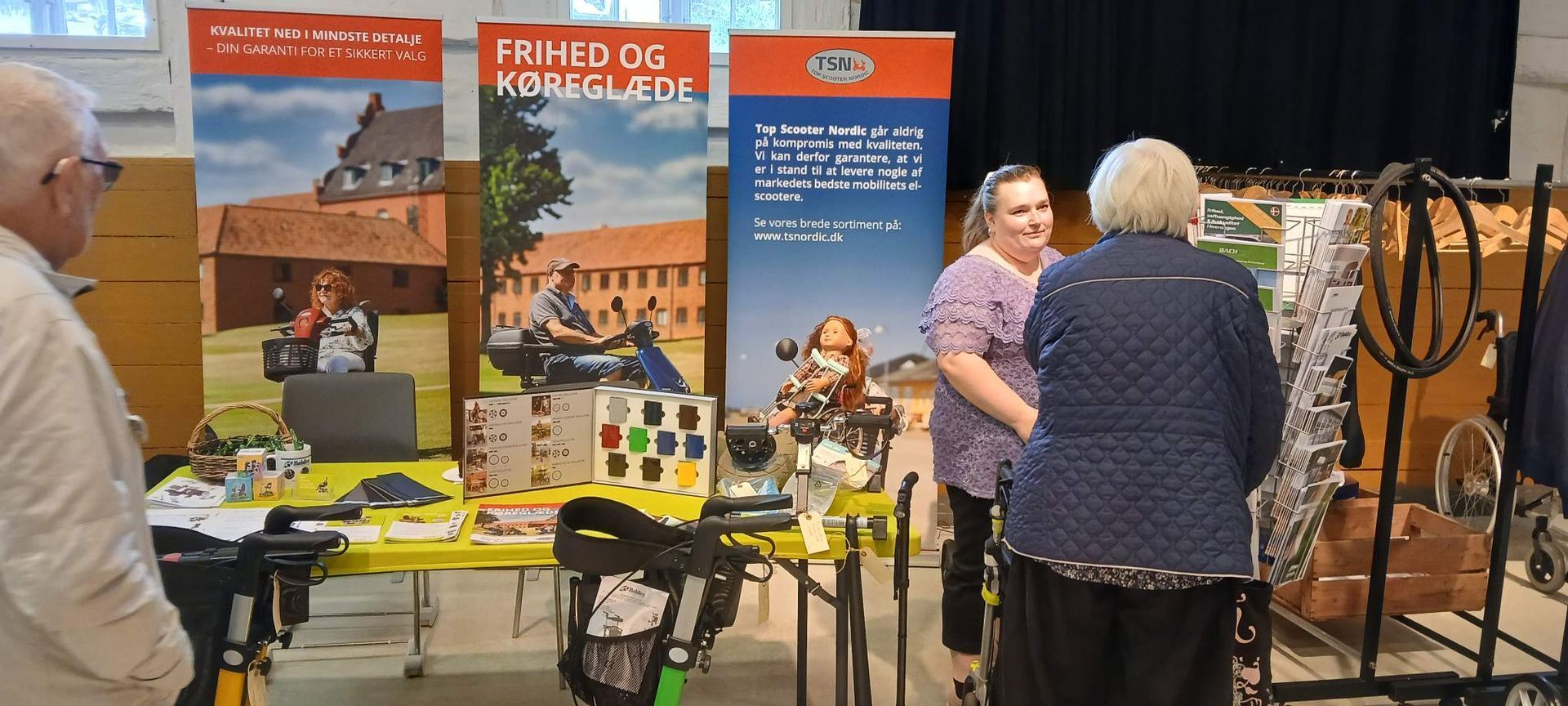 Sara og kunde ved en messestand med informationsbannere og varer til salg.