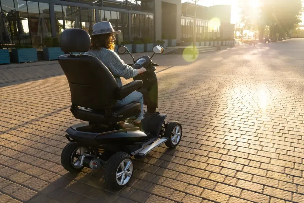 Person i motoriseret scooter på en murstenssti, nær en bygning, baggrundsbelyst af solen.