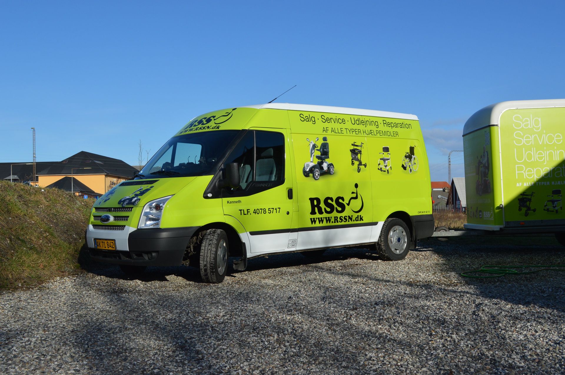 Grøn og hvid varevogn med RSS-logo parkeret på grus i solrige omgivelser.