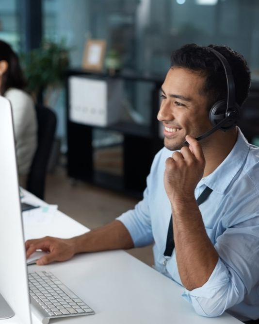 A professional wearing a headset smiles while working on a computer in a bright, modern office.