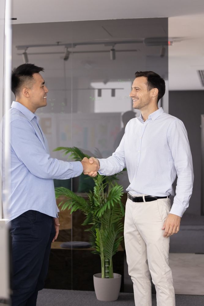 Two people in business casual attire shaking hands while smiling in an office setting with a potted plant.