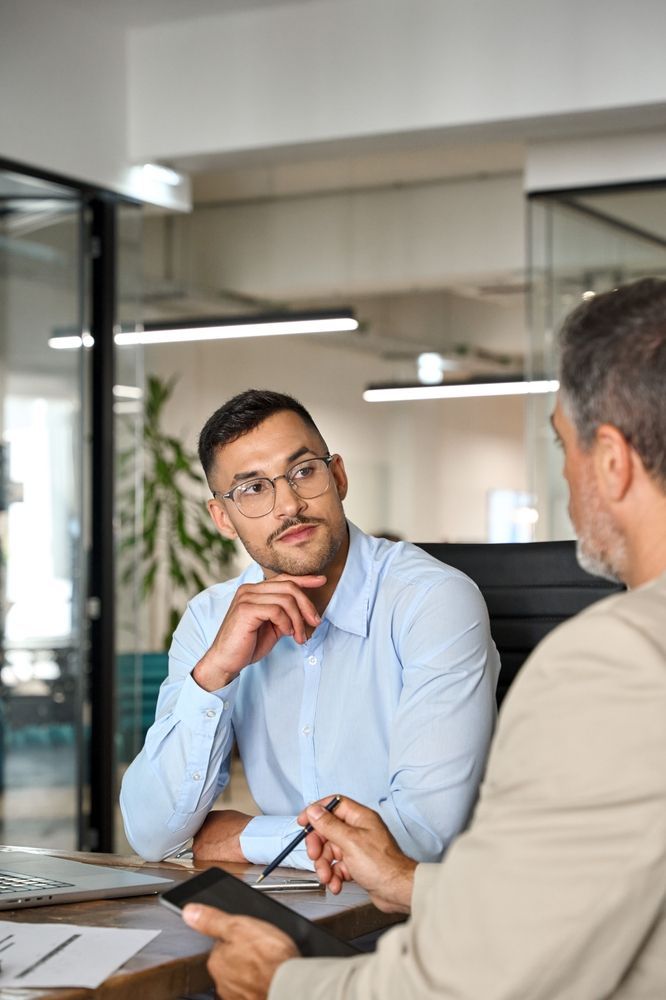 A person in a light blue shirt listens attentively to a colleague holding a tablet during a meeting in a bright office.