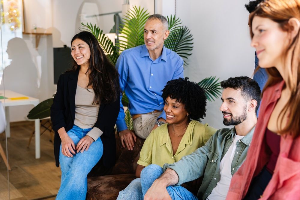 Group of five people seated and smiling in a bright meeting room with plants and a flip chart.