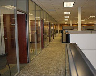 Office hallway with glass-walled offices on the left and cubicles on the right. Beige carpet, fluorescent lights.