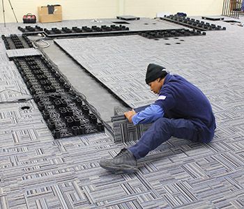 Person installing modular flooring in a large room. Black flooring pieces are being assembled on gray patterned carpet.