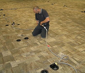 Man kneeling on carpet, connecting cables to floor outlets. Setting: a large room.