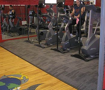 Gym with exercise bikes on a gray carpet floor, people working out. Wooden floor with mural in foreground.
