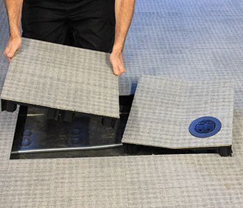 Person removing carpet tile to access a floor outlet. Black plastic frame sits recessed in the carpet.