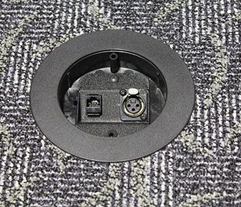 Round black floor port with an Ethernet port and XLR connector, set in patterned gray carpet.