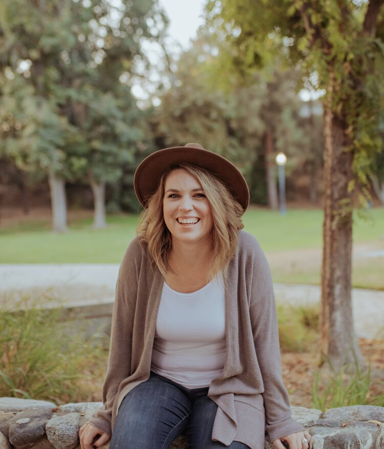 Woman with hat smiles, sitting outside in front of trees, wearing sweater and jeans.