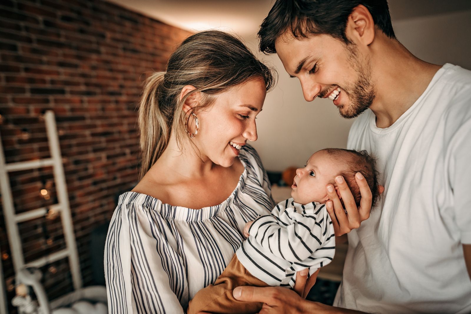 Parents gazing at their newborn baby, smiling with joy. Interior setting with a brick wall.