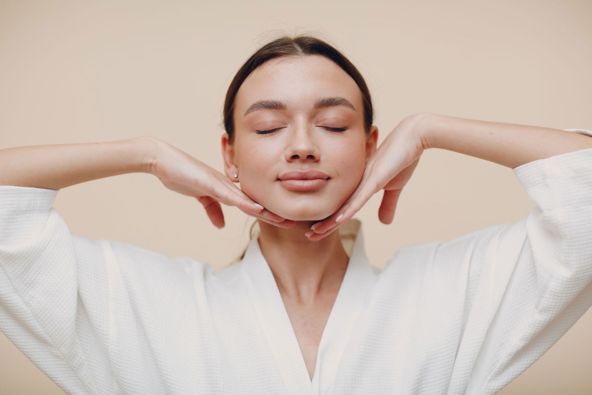 A woman in a bathrobe is making a face massage with her hands.