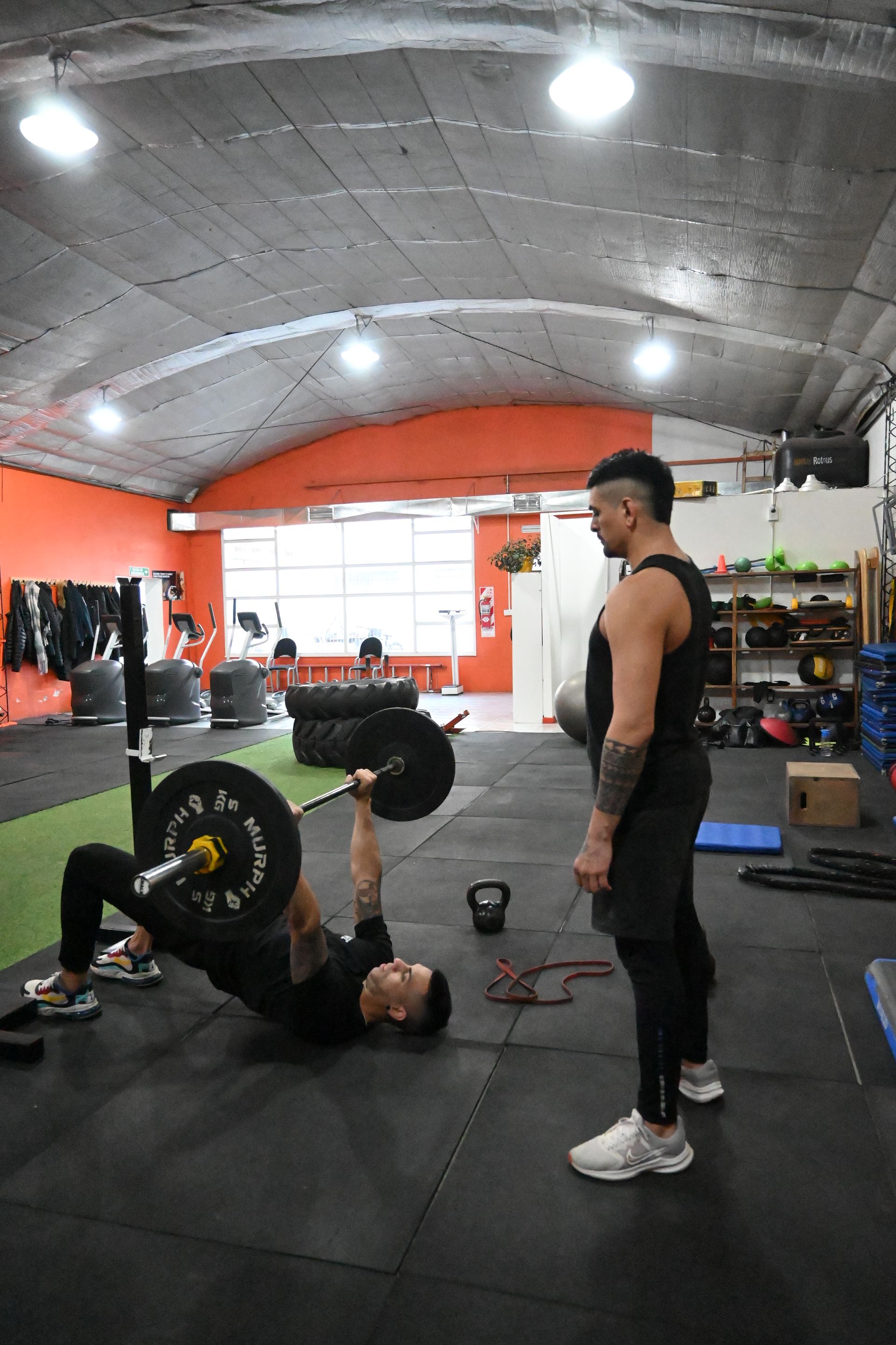 Un hombre está levantando una barra en un gimnasio mientras otro hombre observa.