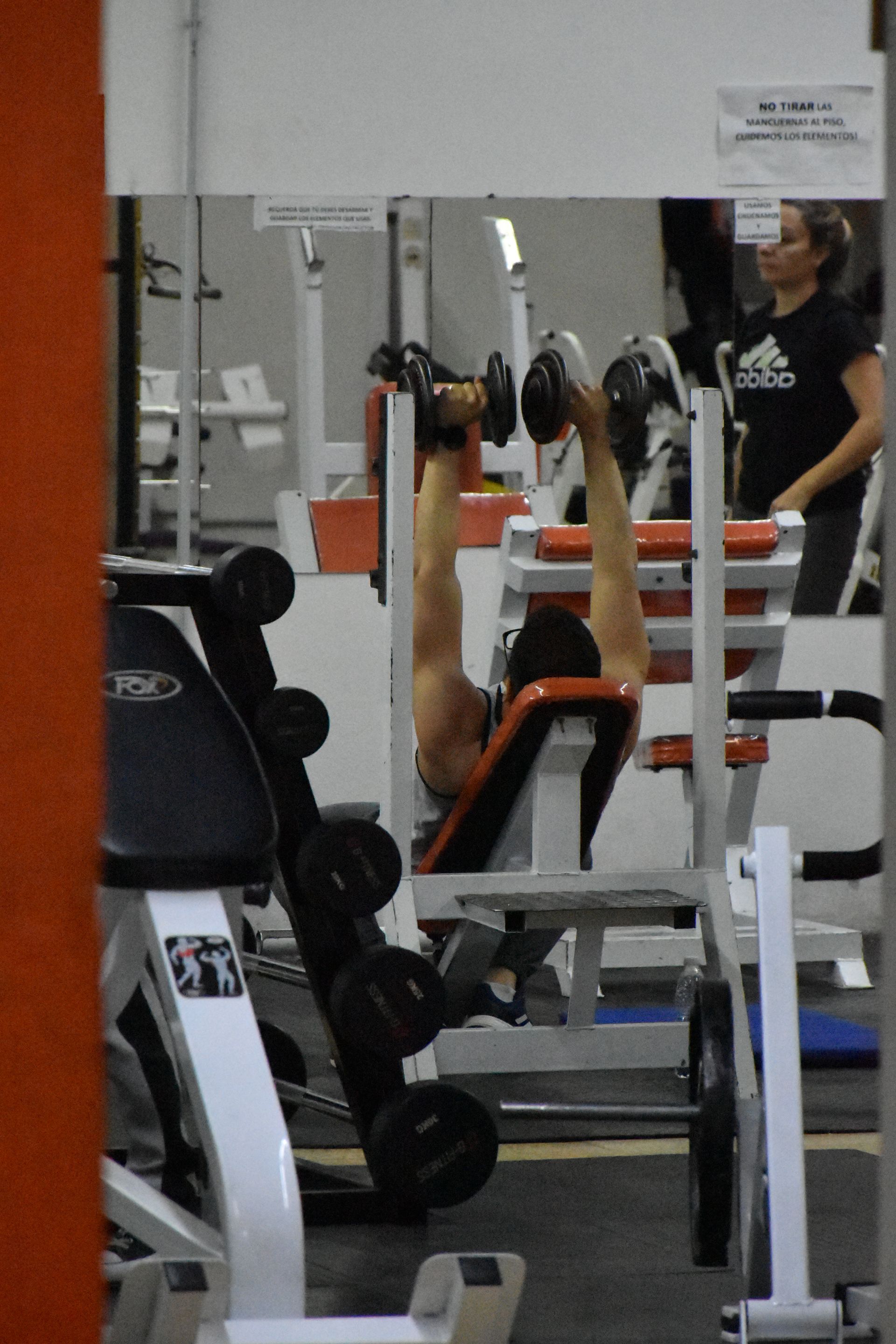 Un hombre está levantando mancuernas en un gimnasio con una camiseta adidas.
