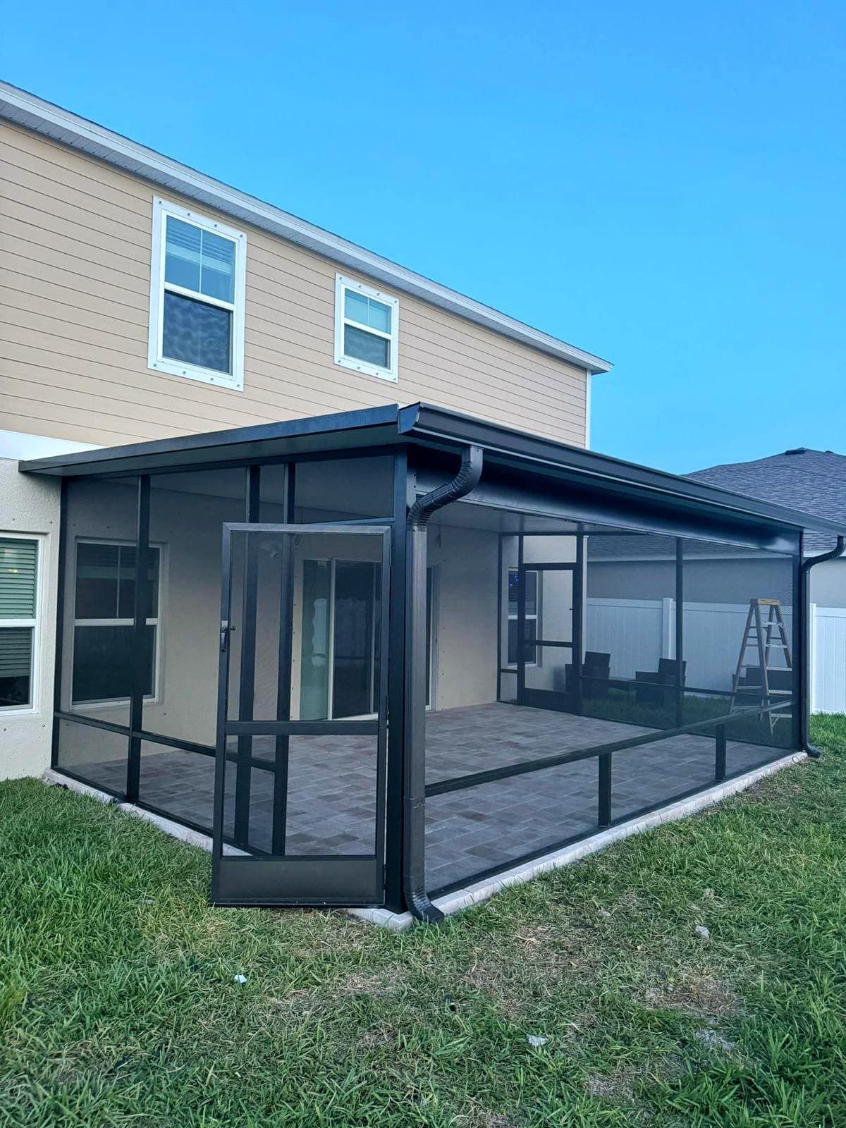 Screened patio addition with dark frame, brick pavers, and door, attached to a beige house, on a sunny day.
