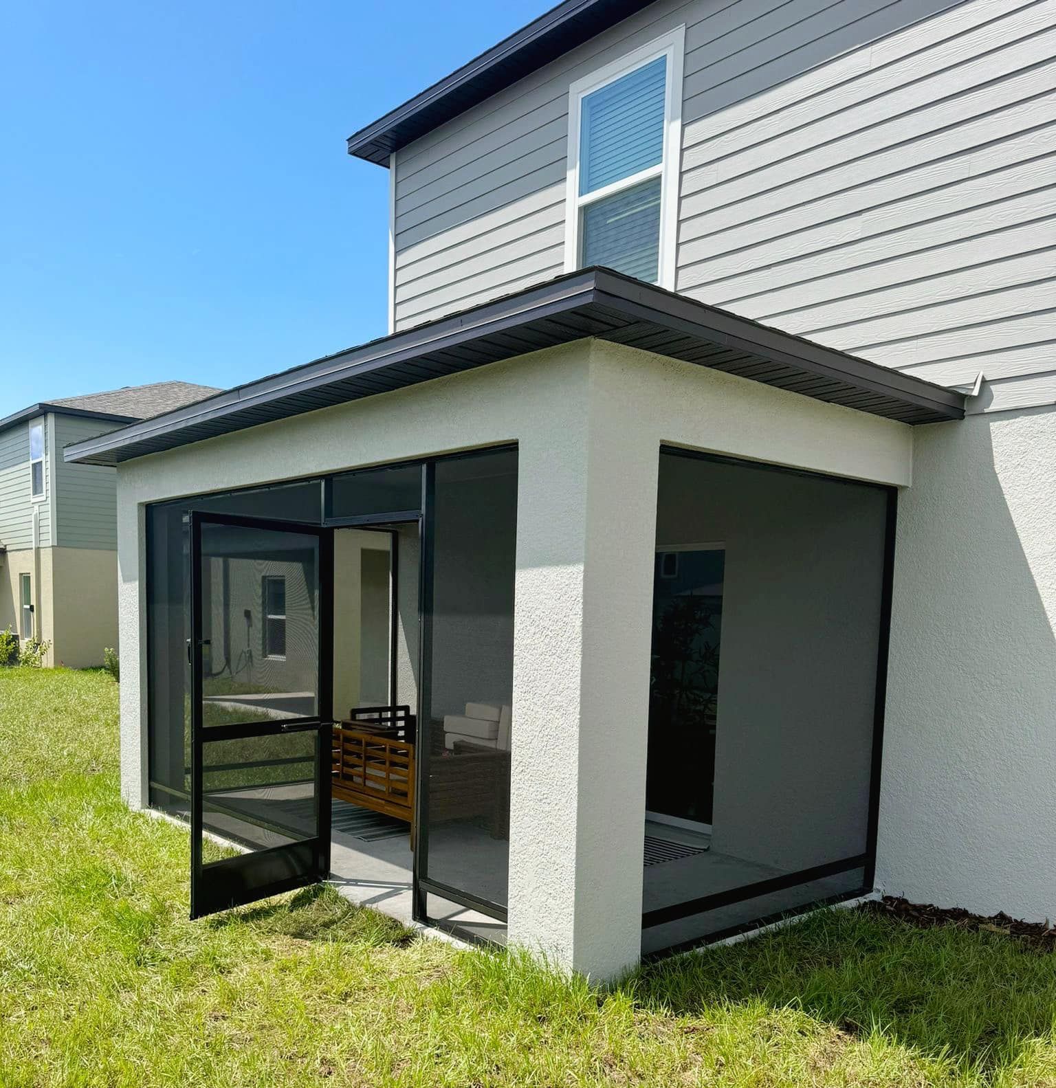 Screened-in patio attached to a house with black frames, rain gutters, and a grassy lawn.