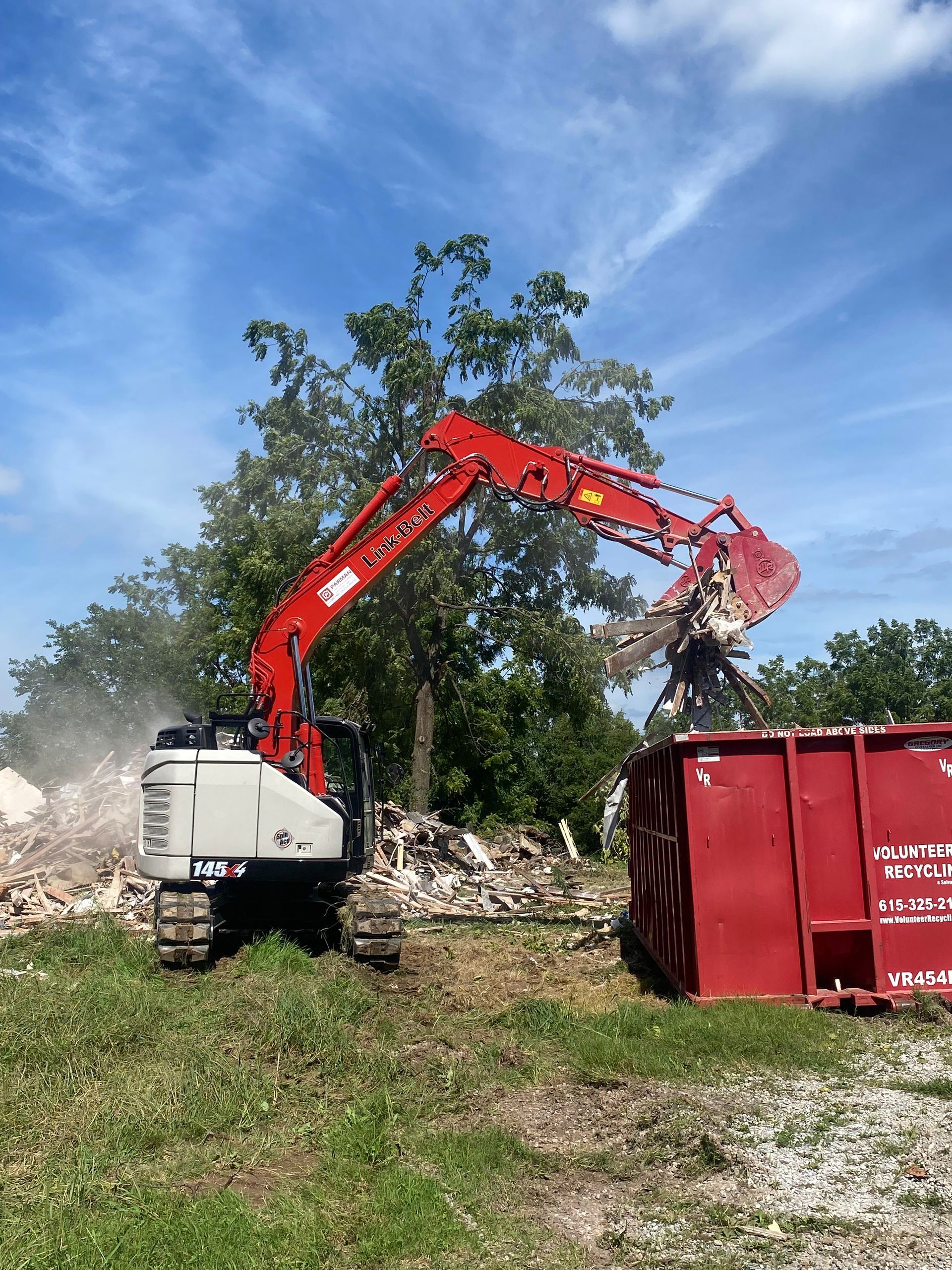 A large excavator is demolishing a building in a field next to a red dumpster.