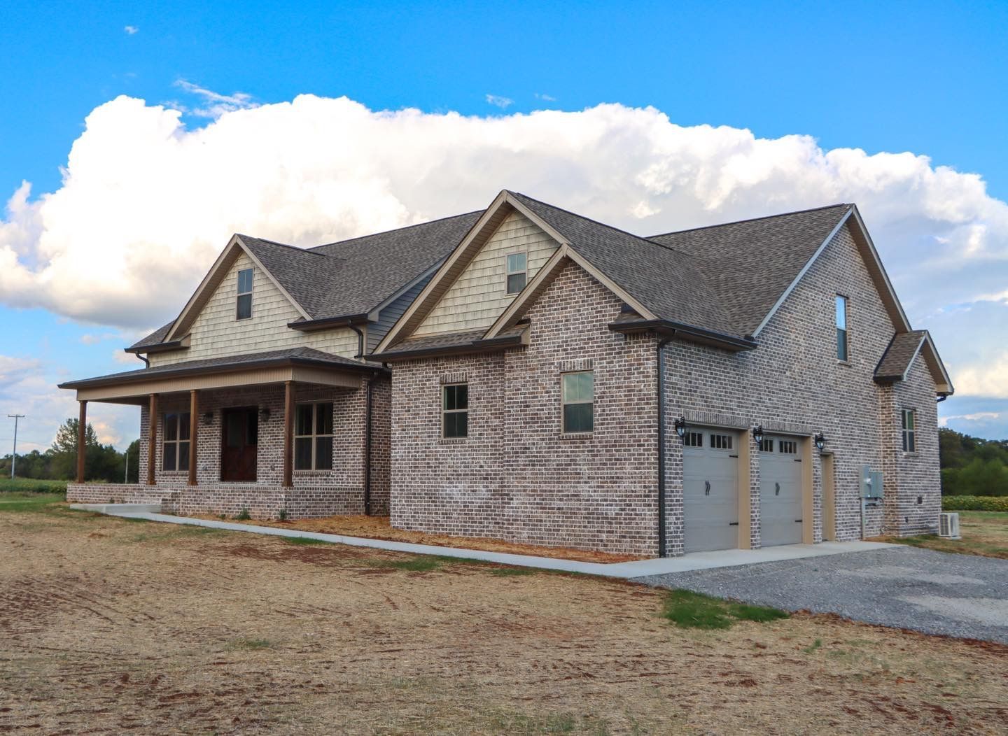 A large brick house with a garage and porch is sitting on top of a dirt field.