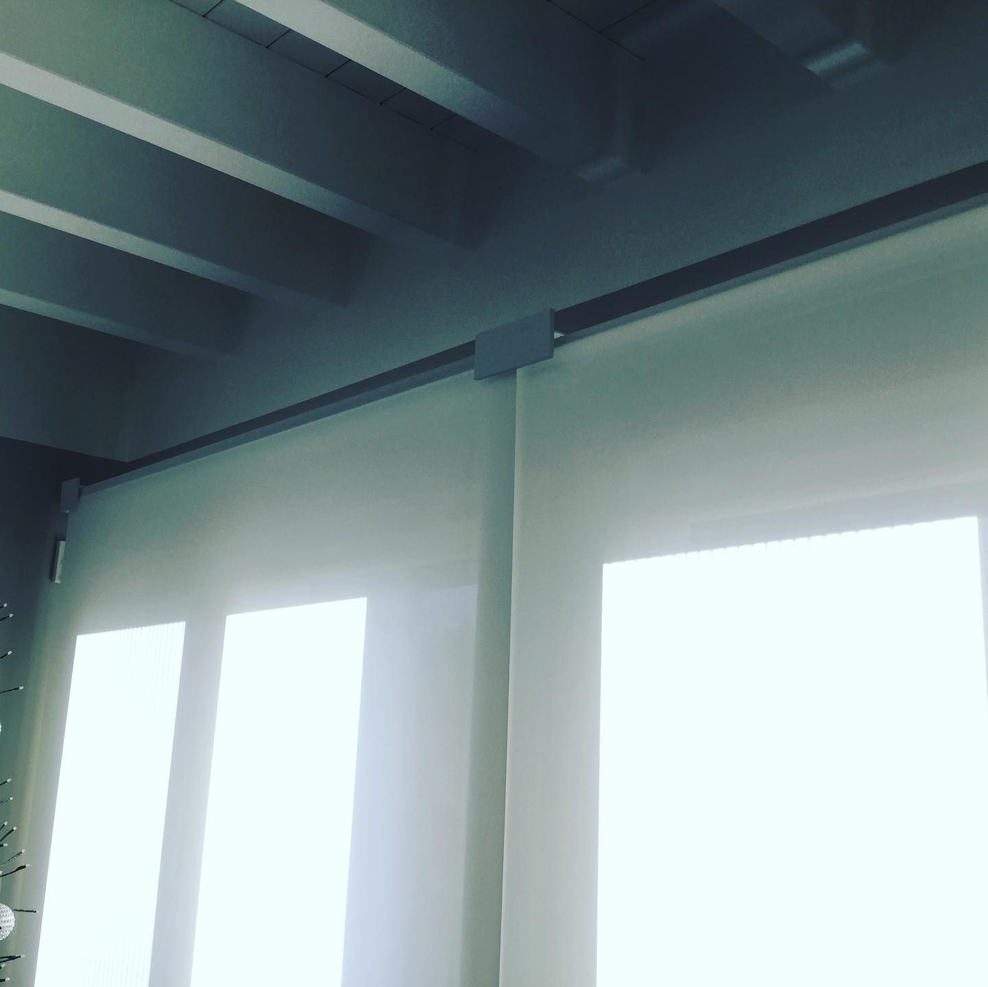 Tende color crema appese a un soffitto con travi grigie. La luce intensa filtra attraverso le tende.