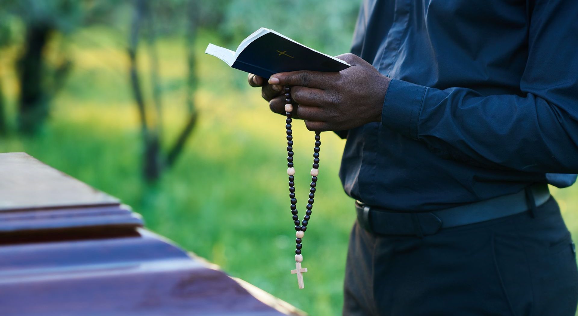 A man is holding a rosary and a bible in front of a coffin.