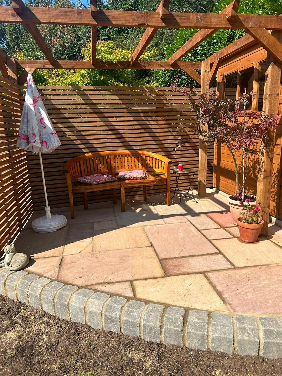 paving garden patio with bench, umbrella, potted flowers, and pergola in sunlight