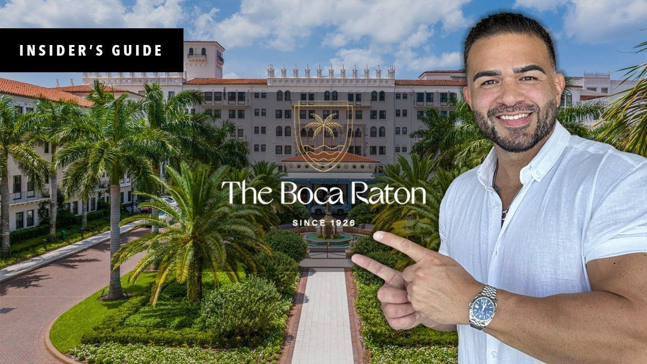 Man pointing in front of The Boca Raton resort. He smiles; trees, lawn, & white building are visible.