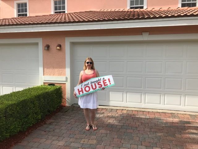 A woman is standing in front of a house holding a sign that says `` i just bought a house ''.
