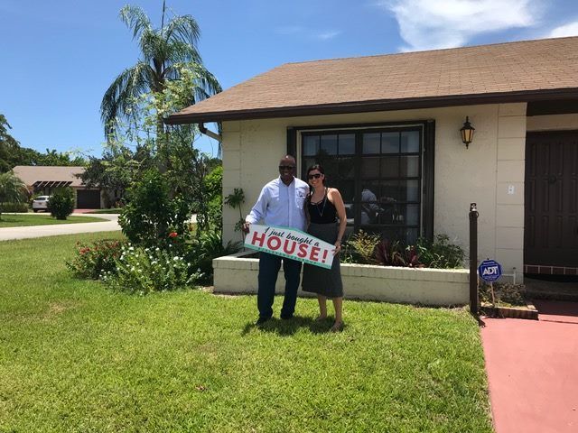 A man and woman are standing in front of a house holding a house sign.