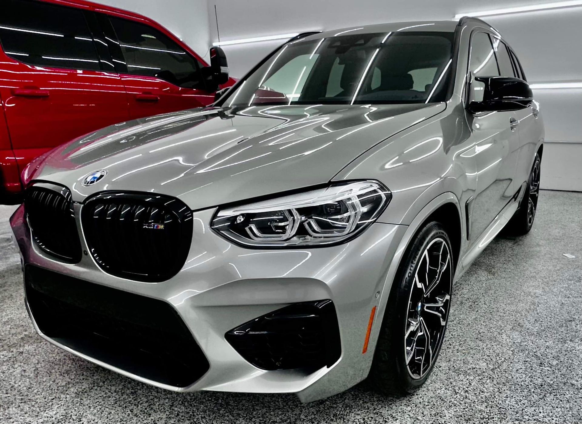 A silver bmw x3 m is parked in a garage next to a red truck.