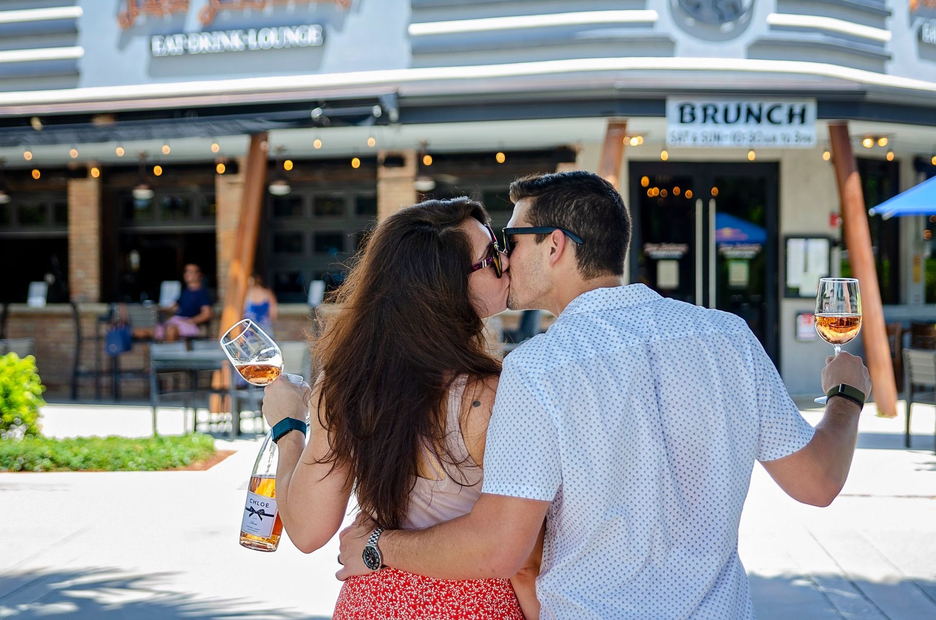 A man and a woman are kissing while holding wine glasses in front of a restaurant.