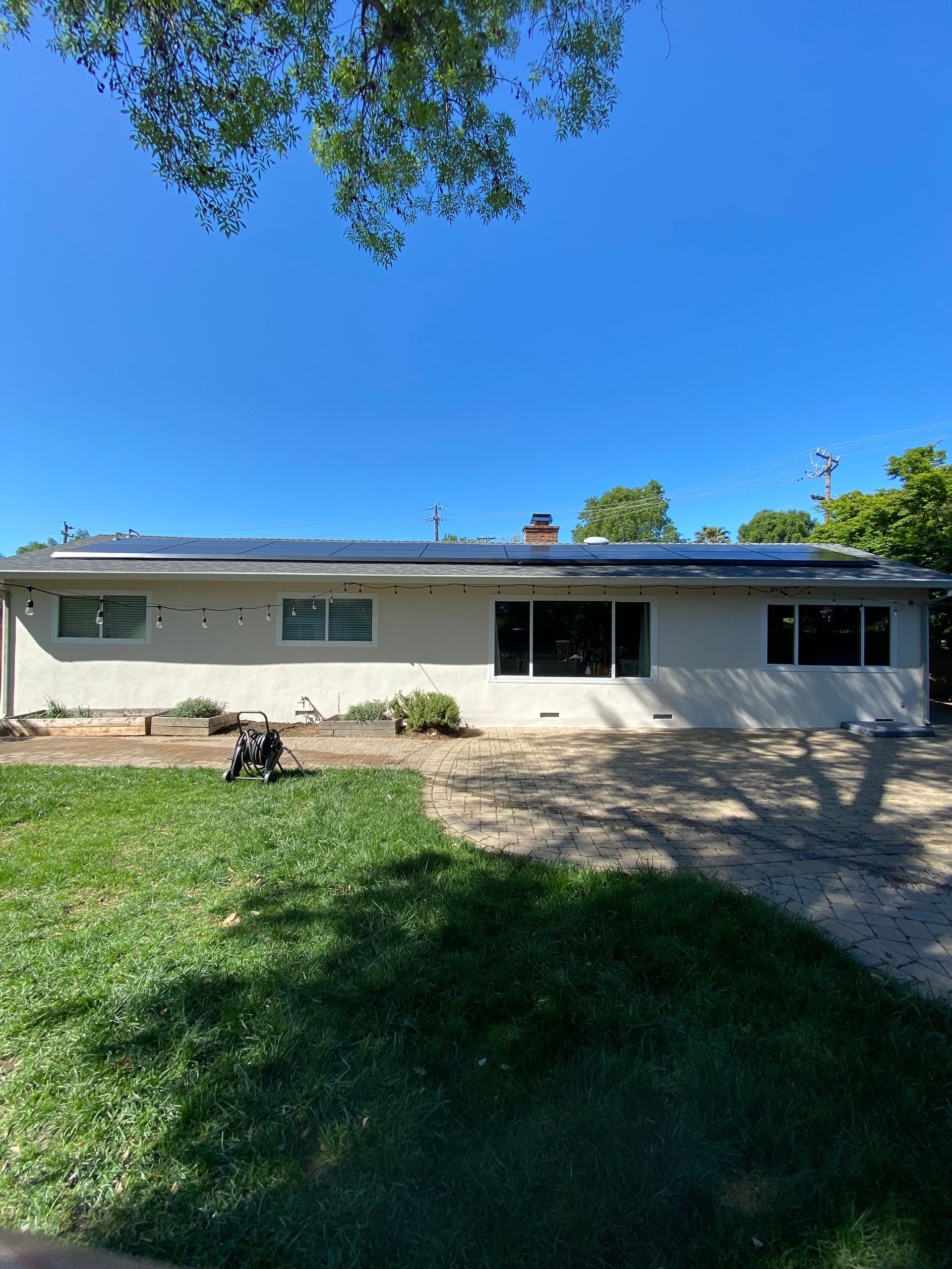 A single-story, light-colored house with a dark roof and solar panels under a clear blue sky, viewed from a grassy yard.