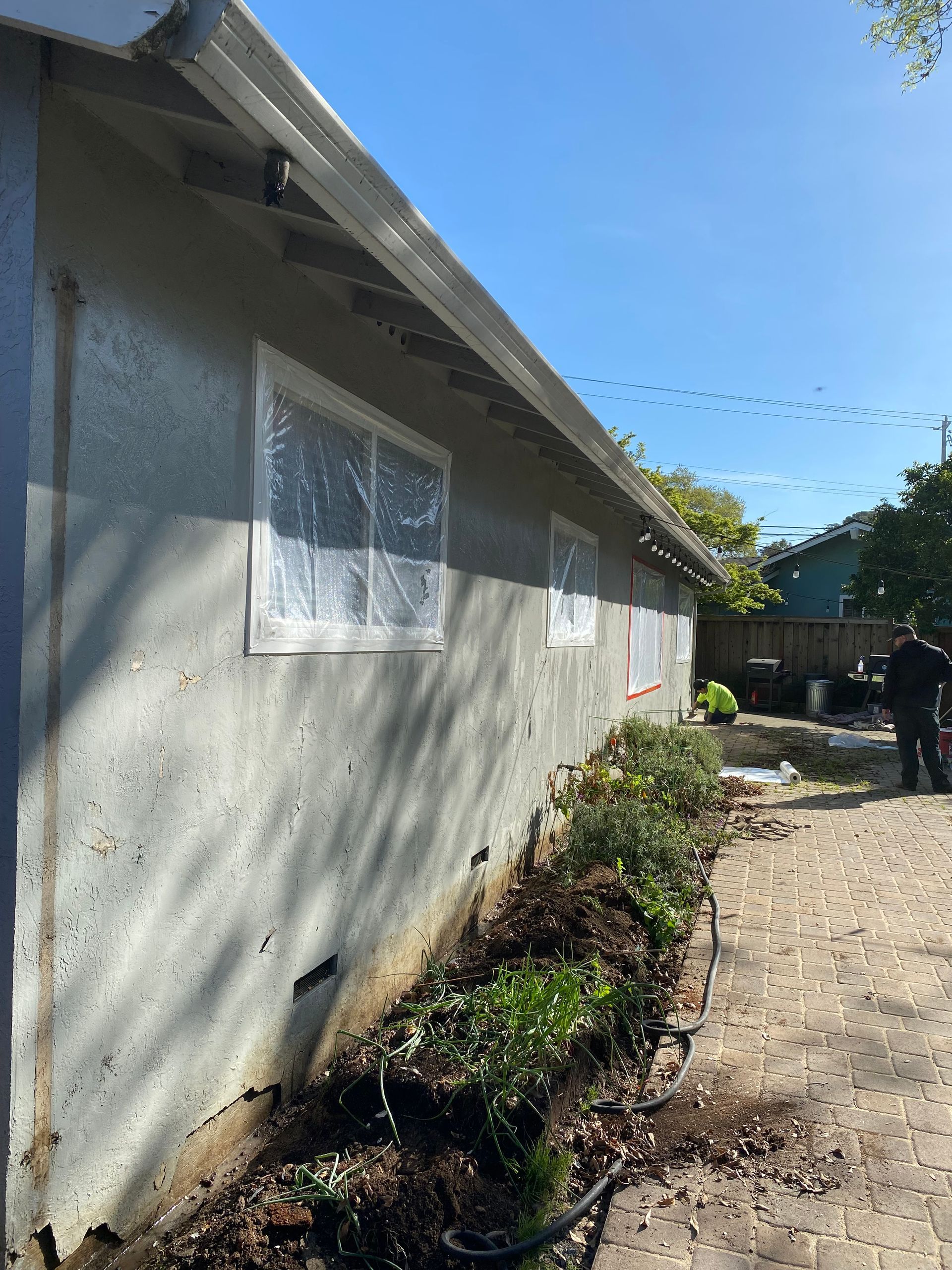 A side view of a house exterior with plastic-covered windows, textured light-colored stucco, and a garden bed below.