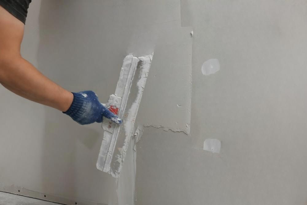 A person wearing a blue work glove uses a taping knife to apply joint compound to a drywall seam on a gray wall.