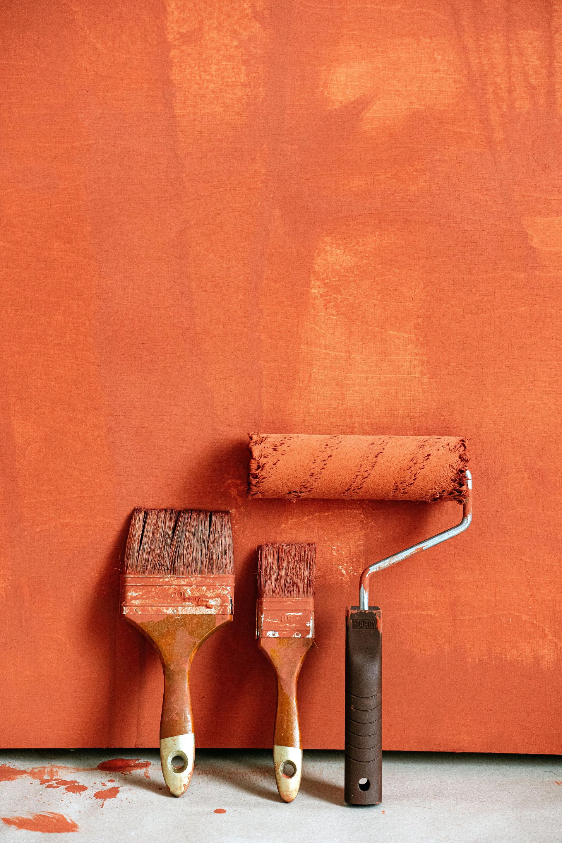 Two paintbrushes and a paint roller covered in orange paint resting against a wall painted in the same orange color.