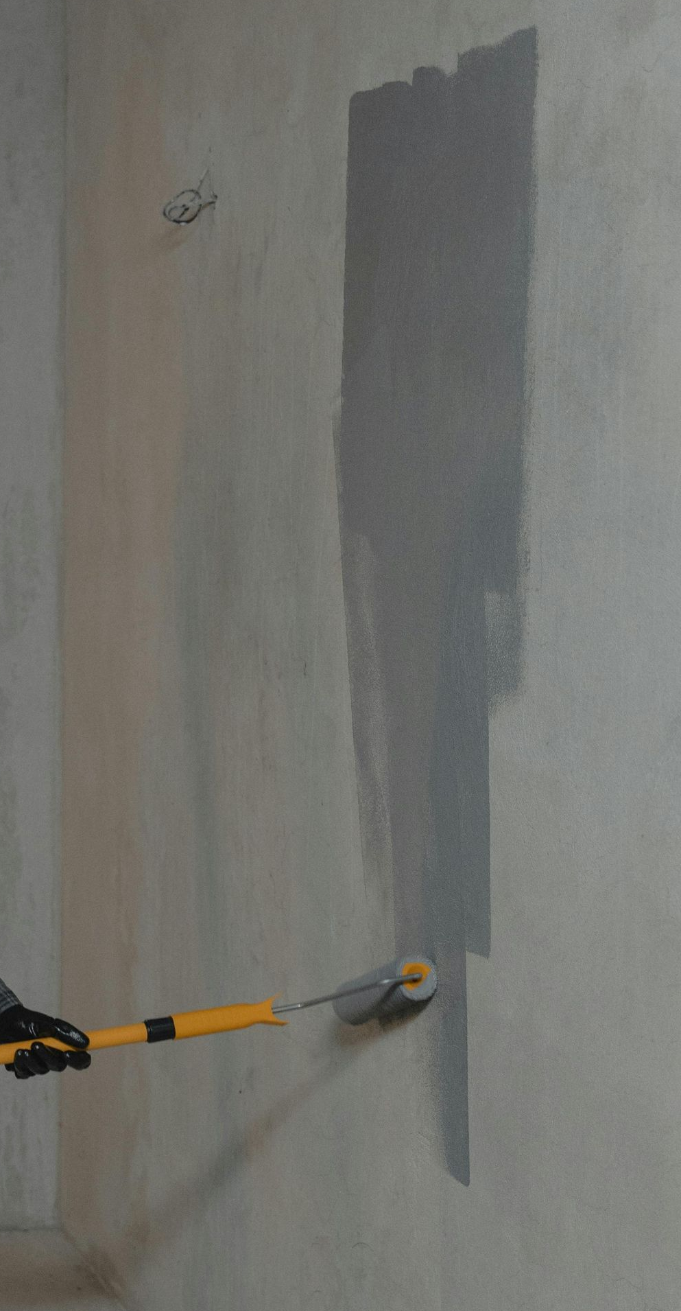 A person using a paint roller with a long handle to apply gray paint to a light-colored wall.