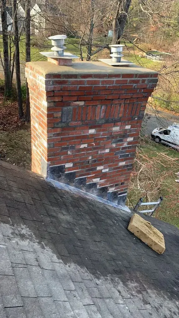 Brick chimney on a dark shingled roof. Brick deterioration visible, with metal caps at the top.