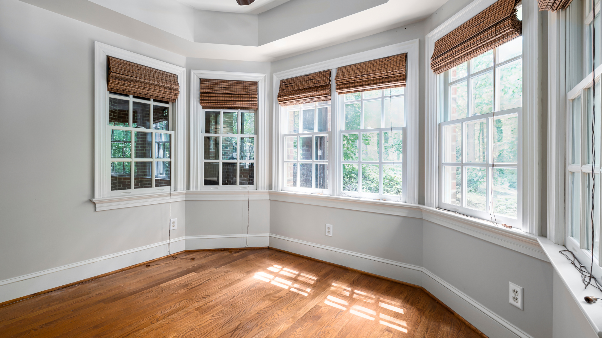 Sunlit interior room with wooden floors, several windows, and woven shades.