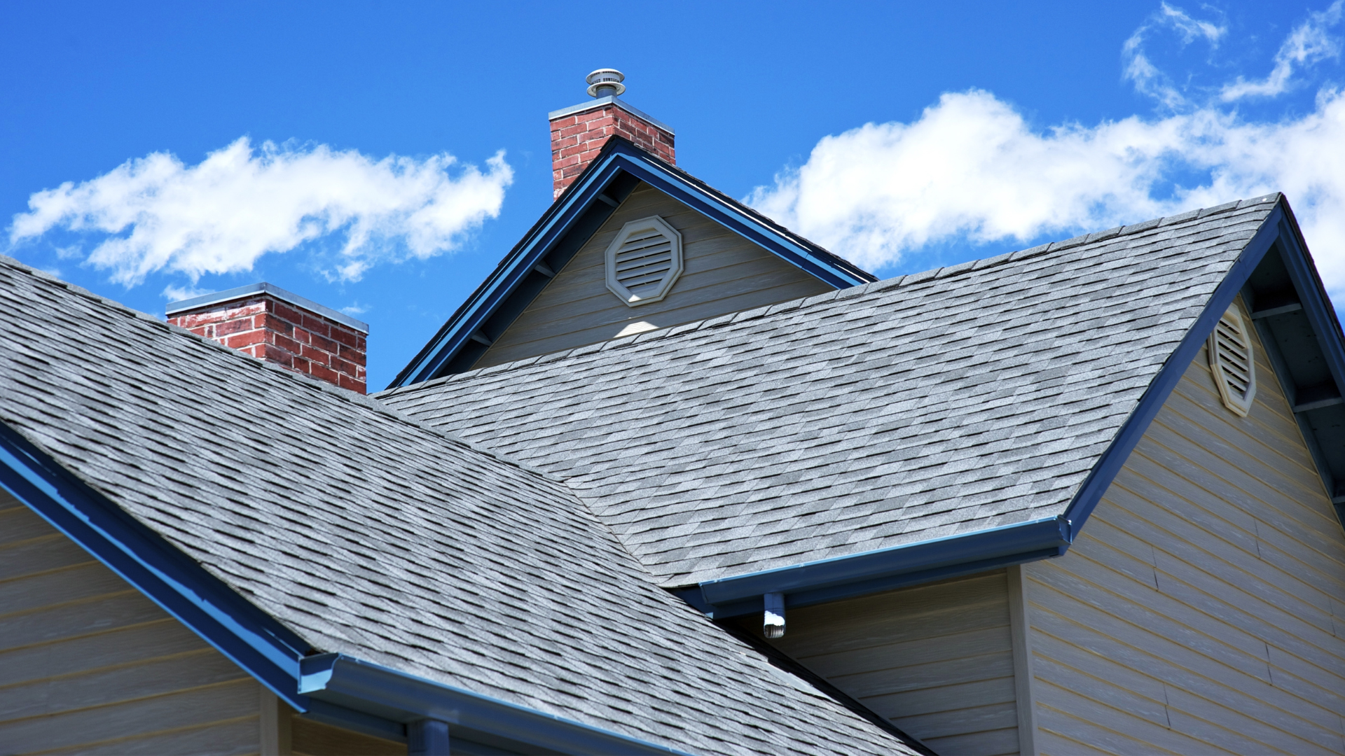 Gray shingled rooftops with blue trim against a light brown house, brick chimneys, and a blue sky.
