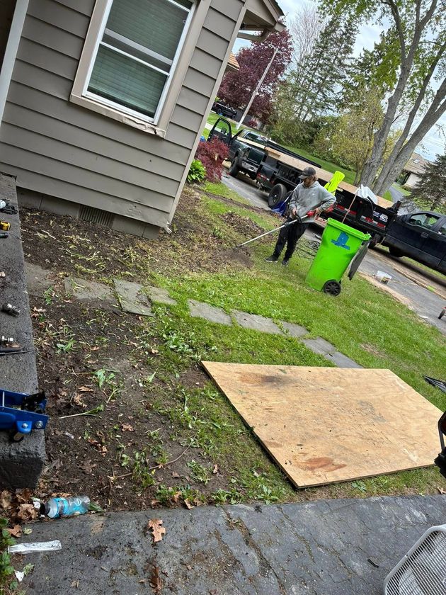 Workers near a house and sidewalk, with a bright green bin and plywood covering a patch of ground. Workers near a house and sidewalk, with a bright green bin and plywood covering a patch of ground.
