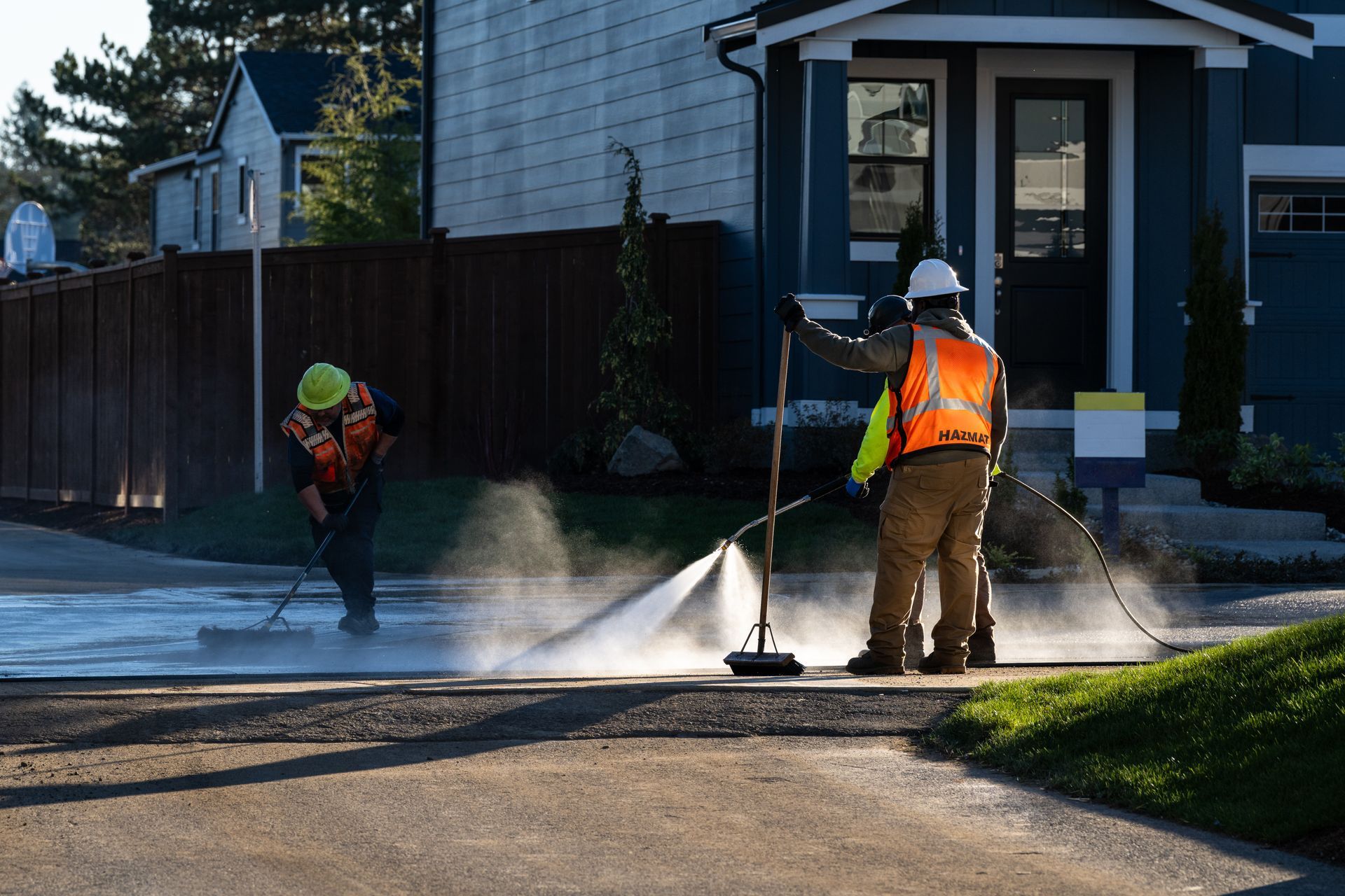 Two workers in hard hats pressure wash a driveway outside a house