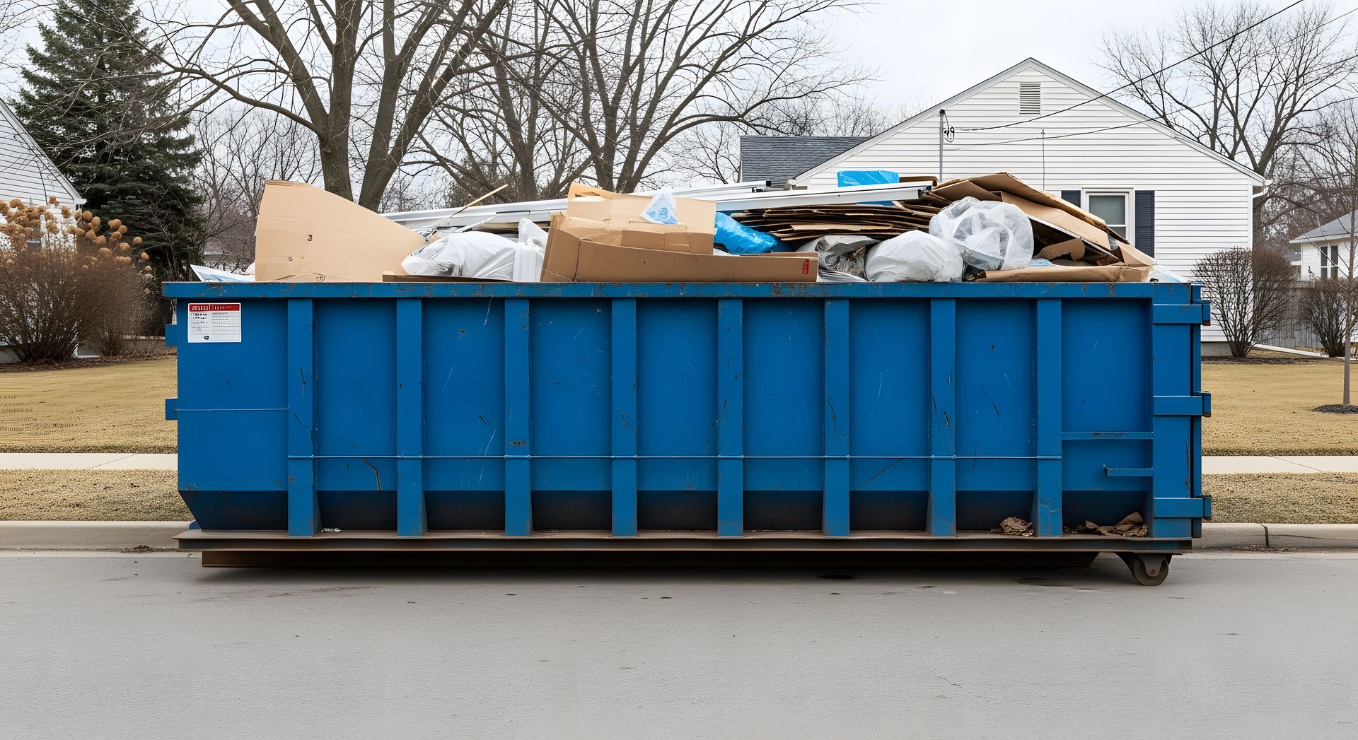 Blue construction dumpster filled with debris and cardboard on a street in front of a house