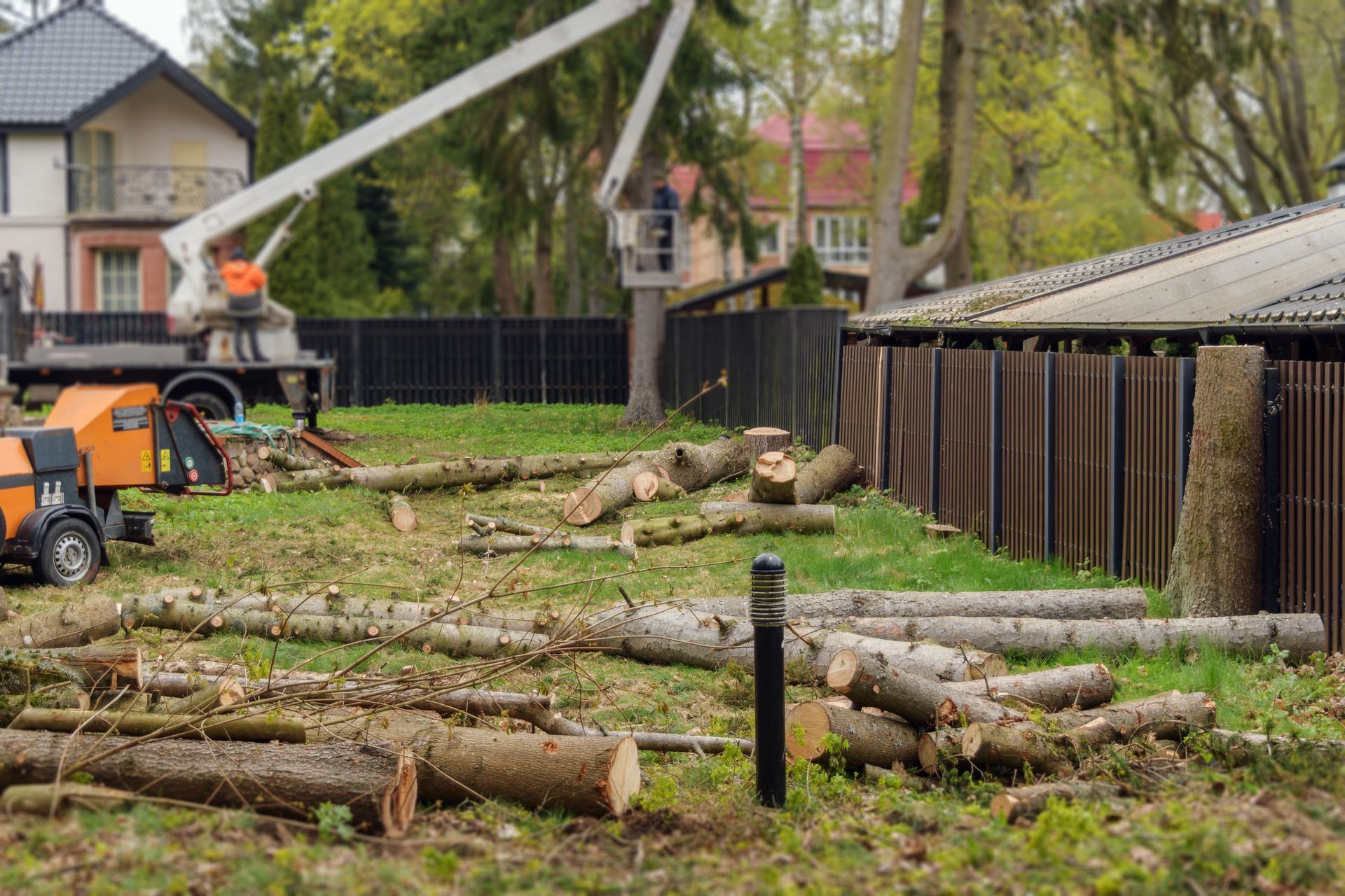 Storm-damaged yard with fallen trees, logs, and a cherry picker beside houses and a fence.