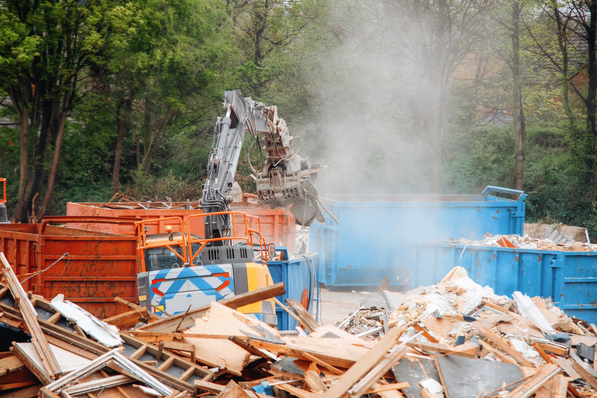 Demolition site with excavator, orange dumpsters, and piles of debris amid dust and trees