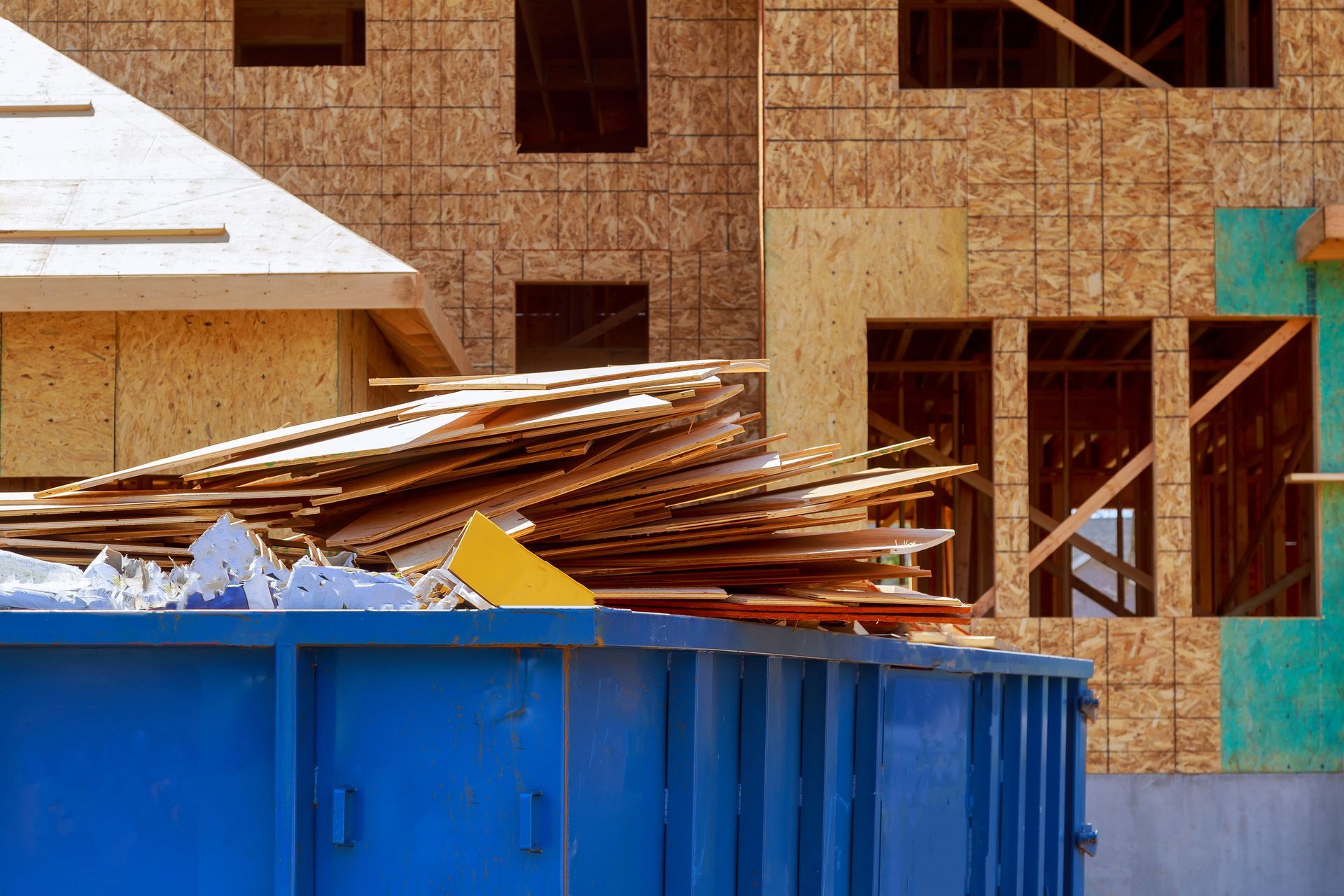 Blue construction dumpster filled with wood debris in front of a building under construction