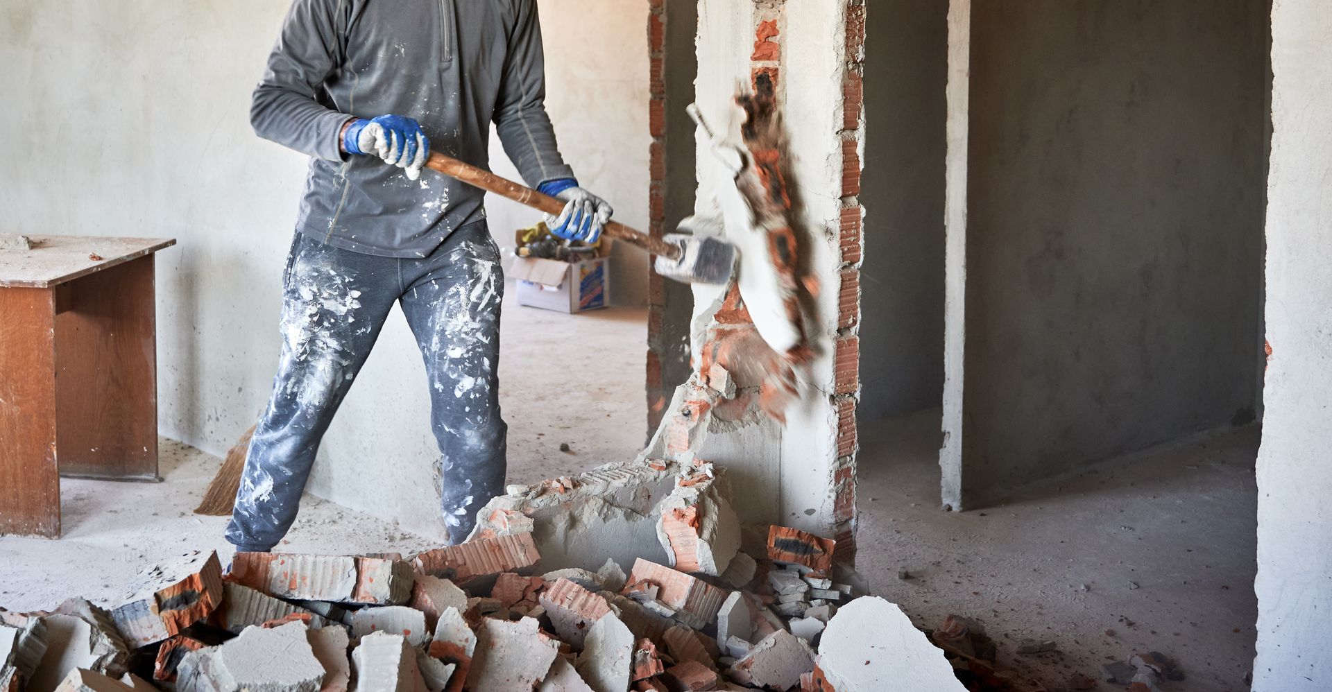 Worker using a jackhammer to break a concrete wall amid rubble inside an unfinished building