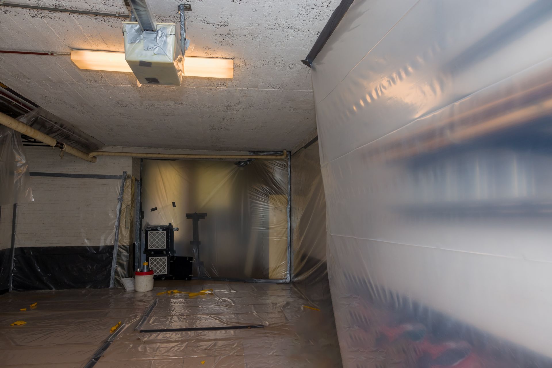 Plastic-covered room under renovation with a ladder, tools, and a ceiling light fixture.