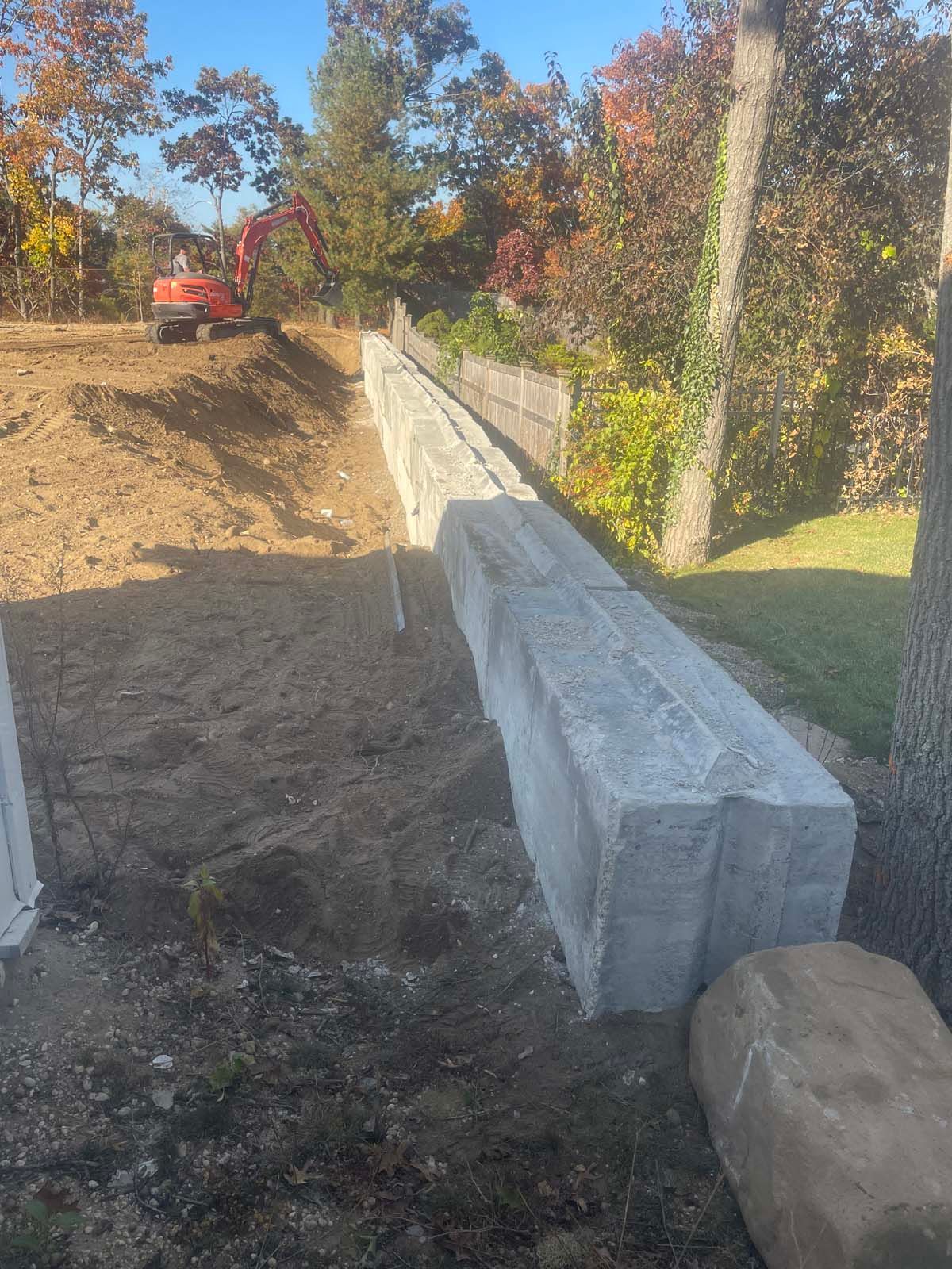 Concrete retaining wall construction site, red excavator, tan dirt, autumn trees.