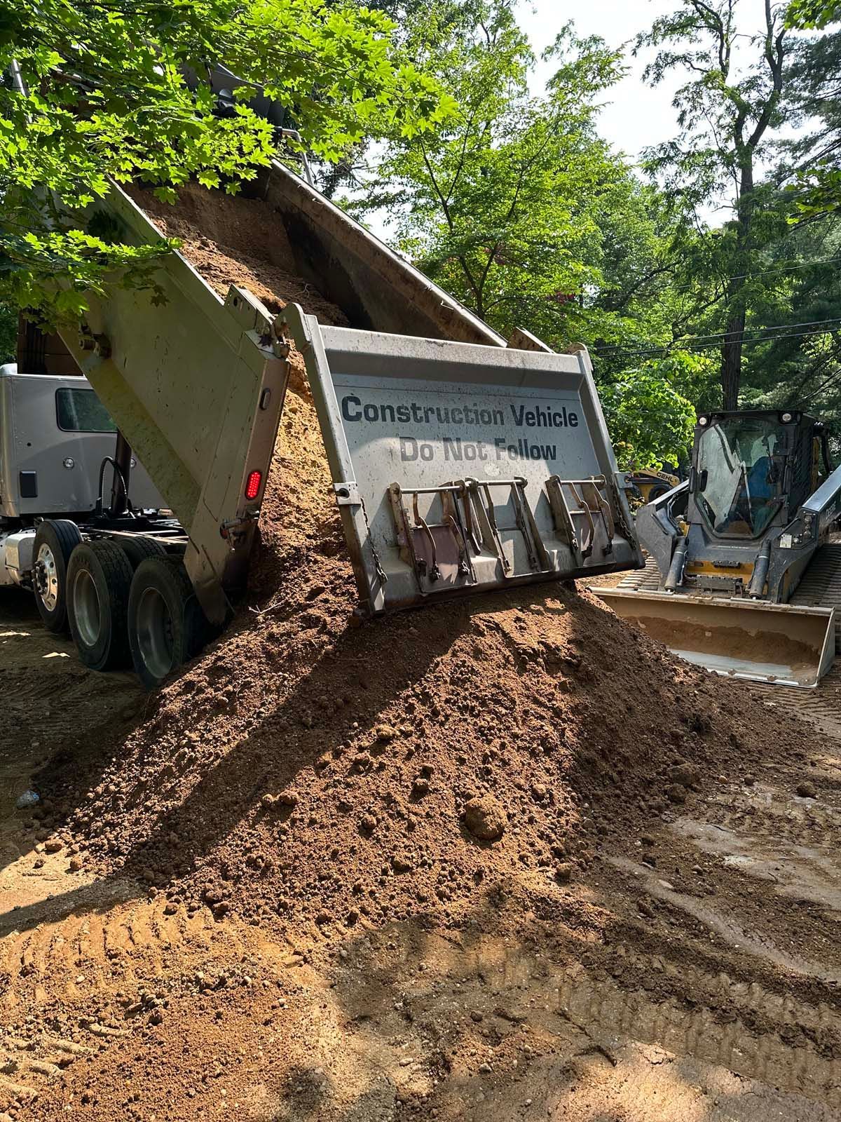 Dump truck unloading soil, construction site. Trees in background, muddy ground.